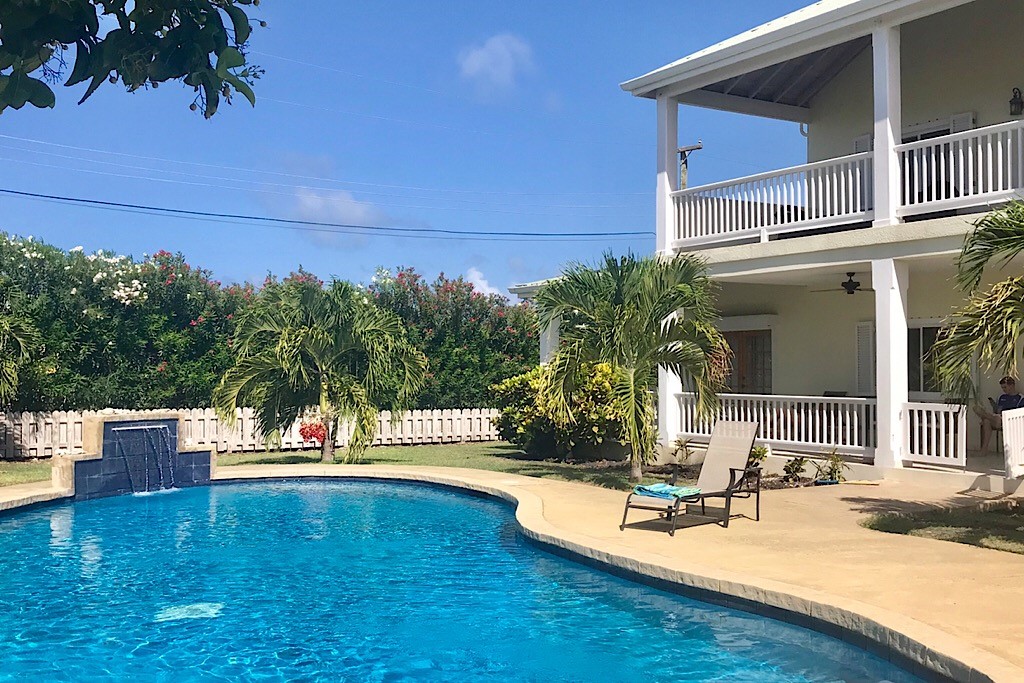 A large swimming pool is surrounded by a spacious patio and tropical foliage. Comfortable lounge chairs are positioned near the water, and the villa features a second story with a broad veranda overlooking the pool area. Bright blue skies complete the inviting atmosphere.