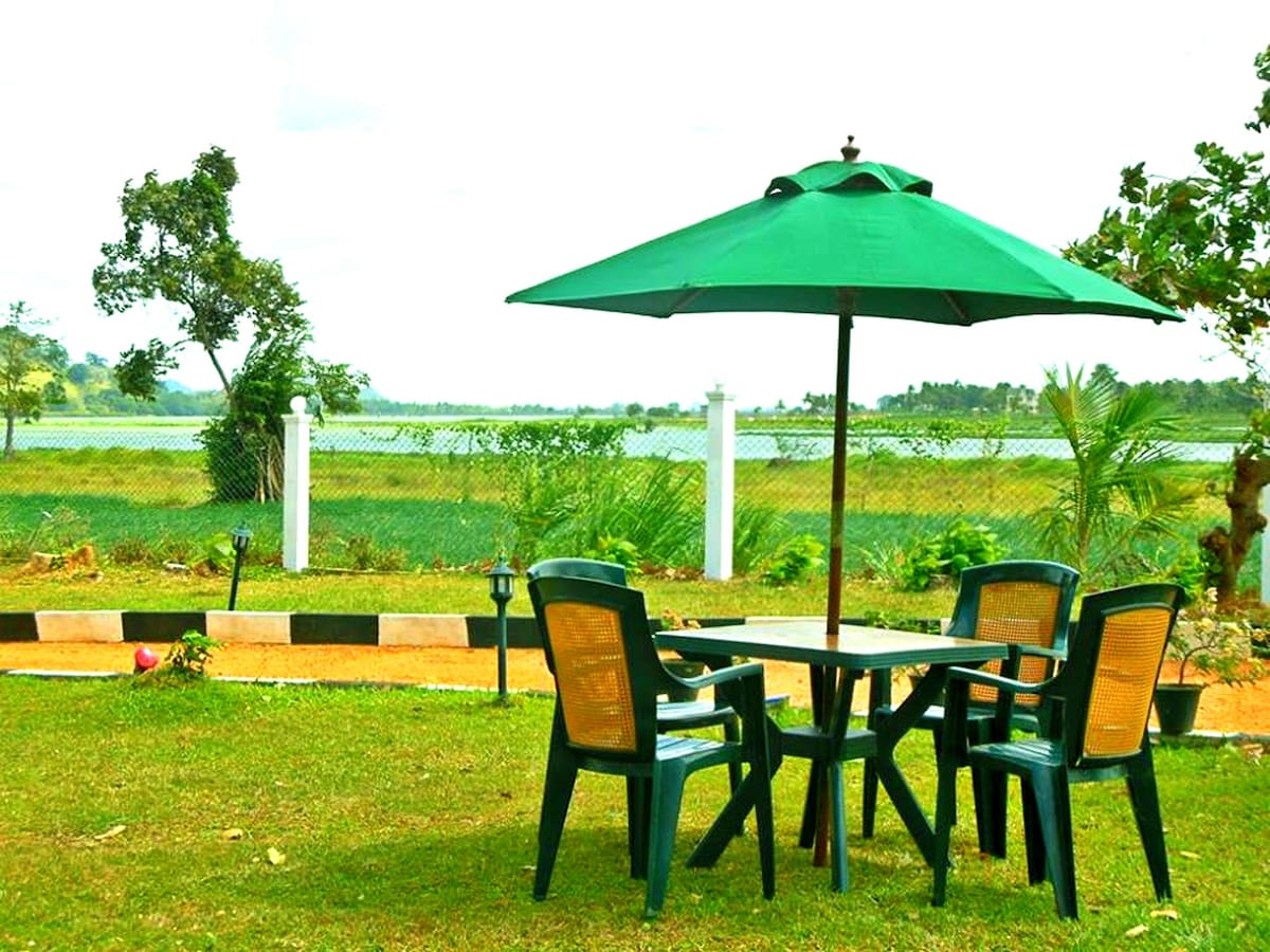 A shaded outdoor seating area is presented with a round table surrounded by four green chairs. A large umbrella offers shade, and lush greenery is visible in the background, along with a serene water view and a well-kept lawn.