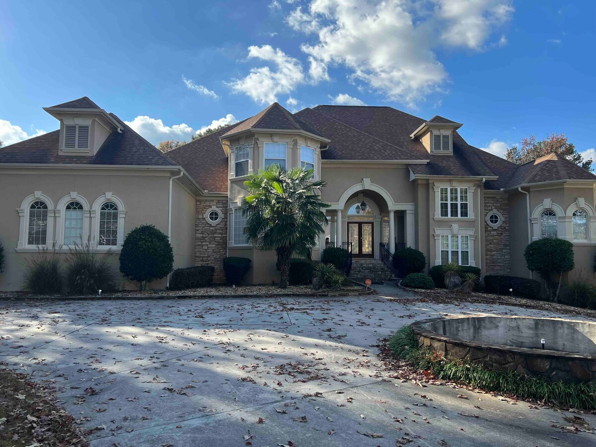 A large, elegant home is presented, featuring a mixture of stone and stucco exterior. Decorative windows adorn the front, while symmetrical landscaping complements the entrance. A circular driveway surrounds a stone fountain, and a clear blue sky is visible above.