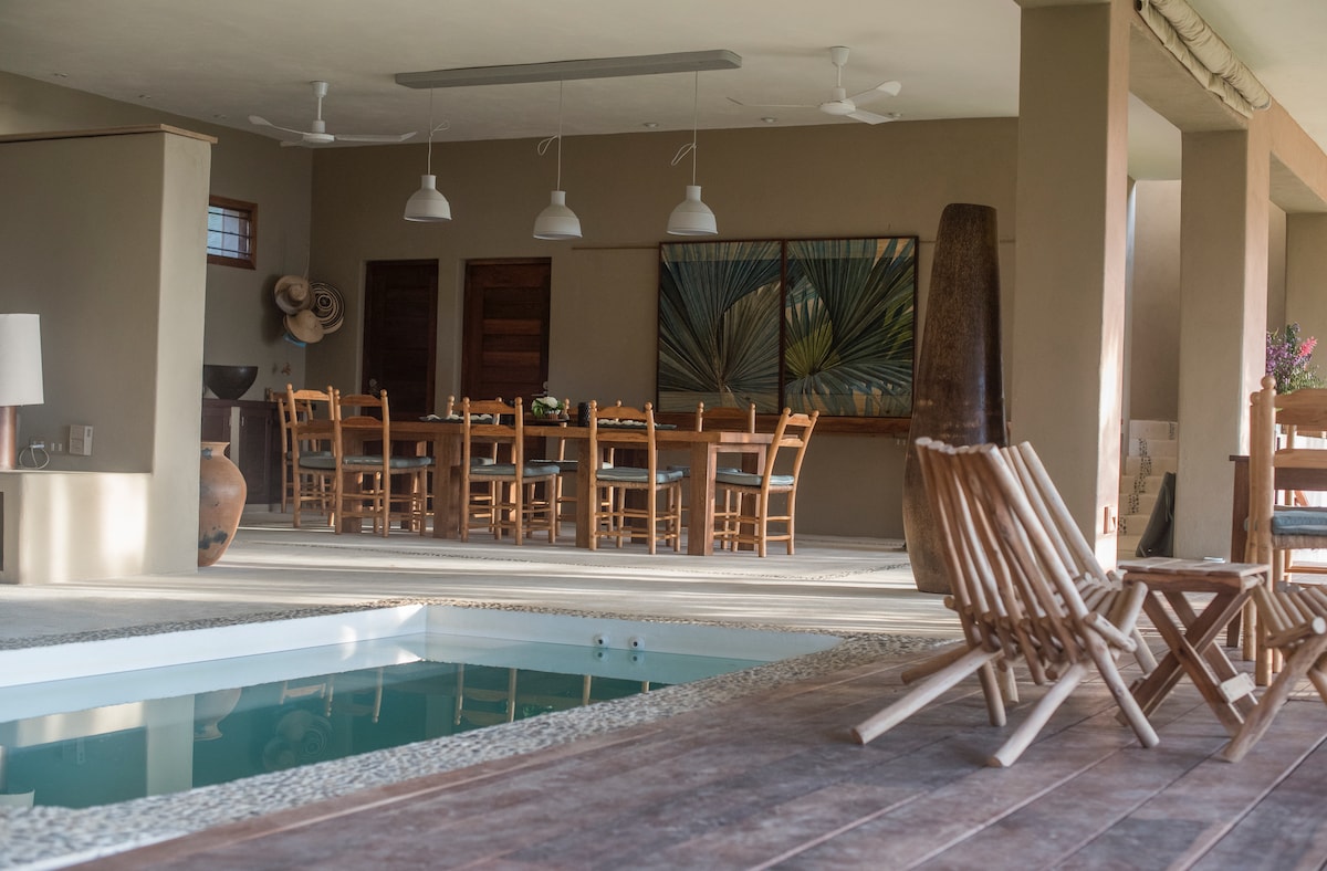 An open living area features a spacious dining table surrounded by wooden chairs, complemented by artful wall decor. Natural light fills the space, reflecting off the indoor pool. Comfortable wooden lounge chairs offer a relaxing spot near the pool's edge.