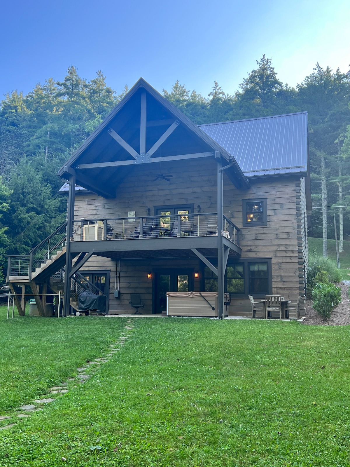 The exterior of a log cabin is showcased, featuring a peaked roof and large deck areas adorned with seating. Lush greenery surrounds the structure, with a stone pathway leading to the entrance. Trees provide a natural backdrop, contributing to a serene environment.