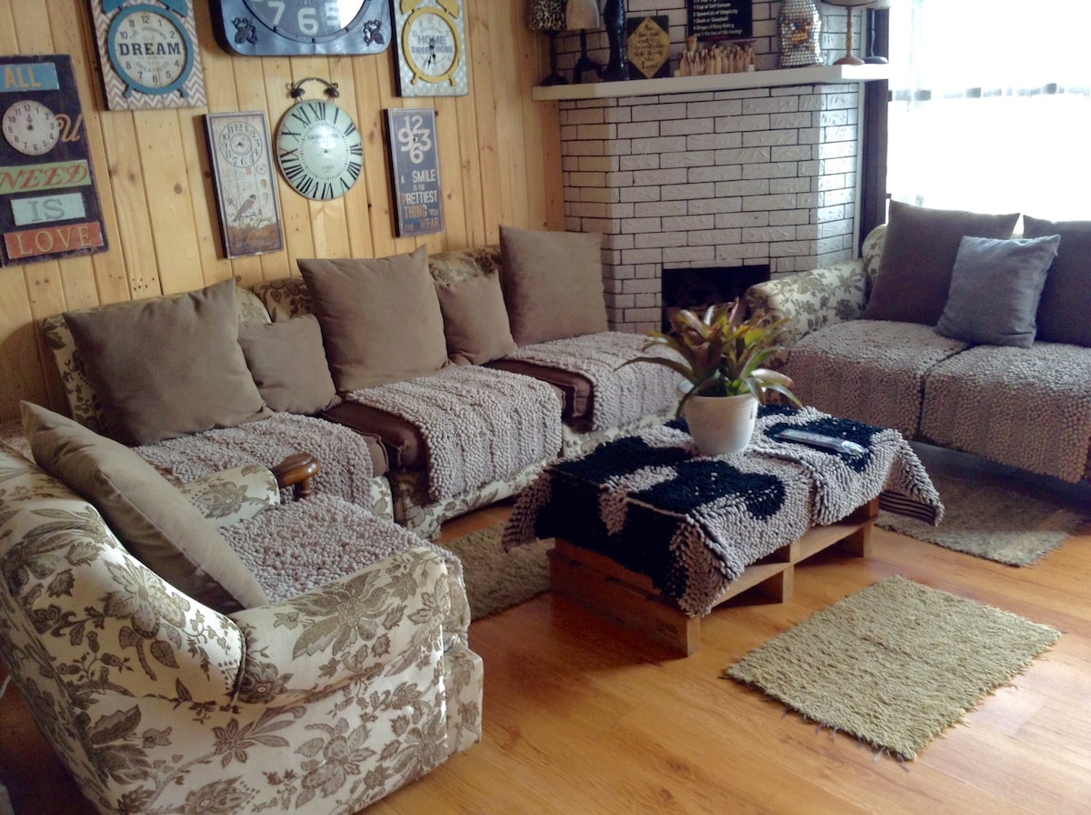 A cozy living room arrangement features a sectional sofa adorned with soft blankets and decorative cushions. A coffee table crafted from wooden pallets sits at the center, complemented by a small potted plant. Warm wood paneling and various wall decor pieces enhance the inviting atmosphere.