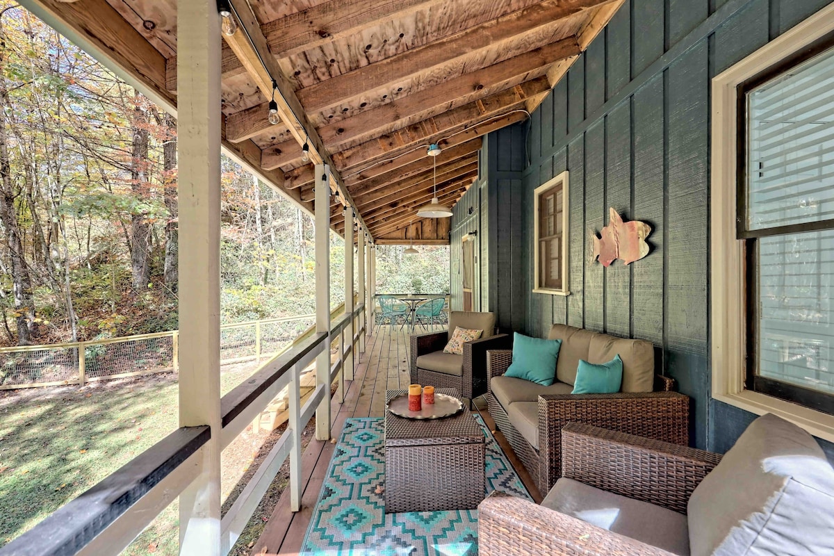 A covered back porch features a conversational seating area with woven furniture, complemented by colorful cushions. A patterned area rug is placed beneath a small table. Soft string lights are visible overhead, creating a warm ambiance in this inviting outdoor space.
