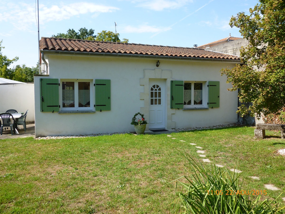 A quaint, renovated single-story house is presented, featuring light-colored walls and green shutters. The entrance is marked by a white door, with a small flowerpot placed nearby. A spacious lawn with stepping stones leads to the home, offering a peaceful ambiance in the countryside.