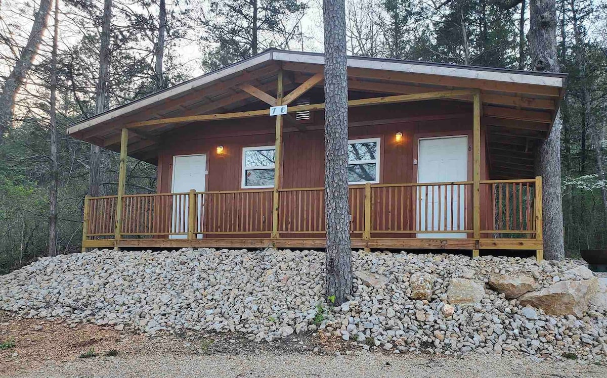A cozy duplex cabin is set on a hillside, surrounded by trees and natural stone landscaping. The structure features a covered porch with wooden railing, providing entry through two white doors. Soft lighting illuminates the cabin's exterior, accentuating the warm wooden finish.