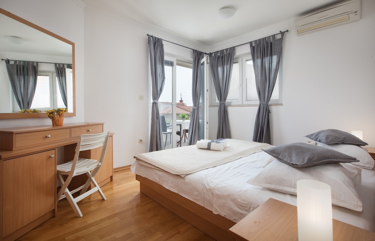 A well-lit bedroom is furnished with a spacious bed featuring light-colored bedding. Two large windows with grey curtains allow natural light to enter, and a wooden dresser with a mirror complements the room. A small desk and chair are positioned nearby, offering a cozy work space.