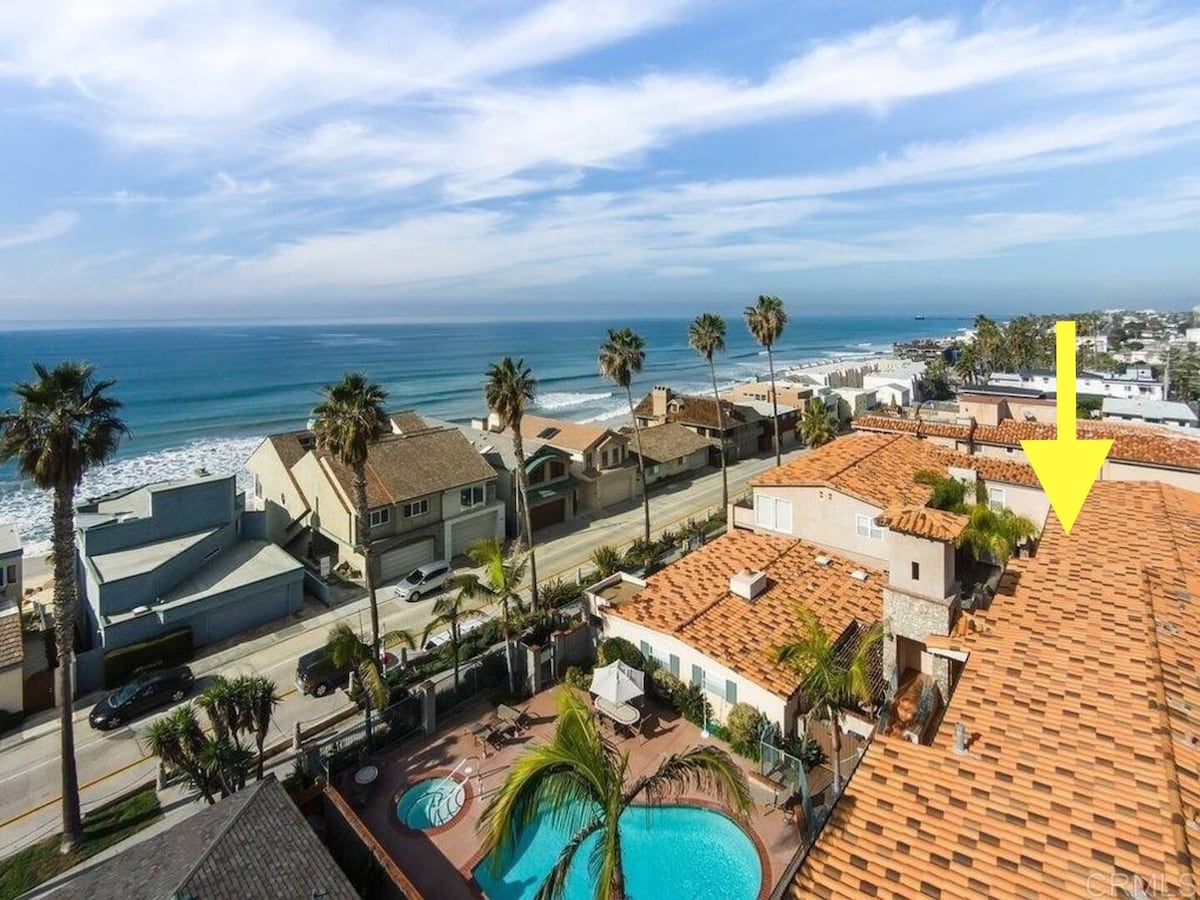An aerial view captures the exterior of the beach house, featuring a vibrant blue ocean and sandy shoreline. The swimming pool below is surrounded by palm trees, with neighboring residential buildings visible, enhancing the coastal ambiance.