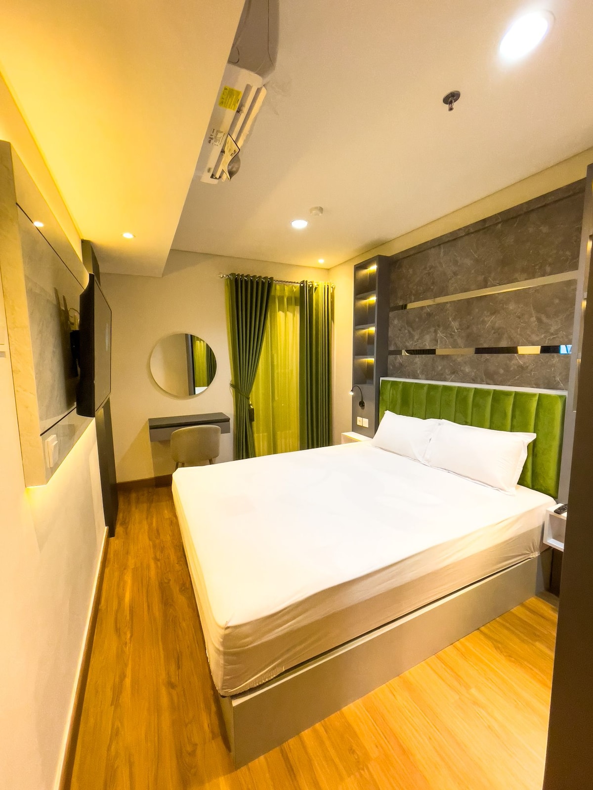 The main bedroom features a spacious bed with a green accent headboard. A smart TV is mounted on the wall, and natural light enters through the large window draped with green curtains. A small desk and chair are positioned near the mirror.