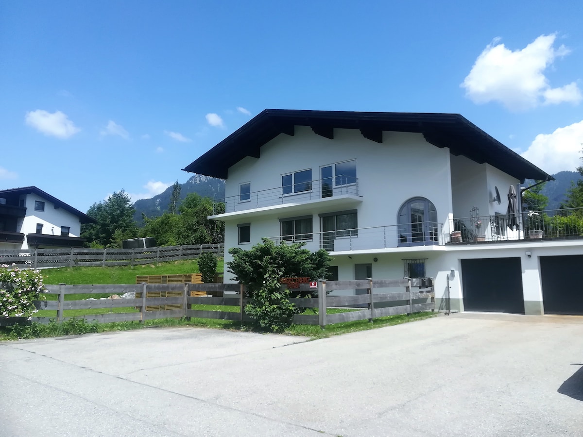 A two-story house is depicted with a sloped roof and large balconies overlooking an outdoor area. The property is surrounded by greenery and features a driveway with ample parking space. A mountain backdrop is visible in the distance under a clear blue sky.