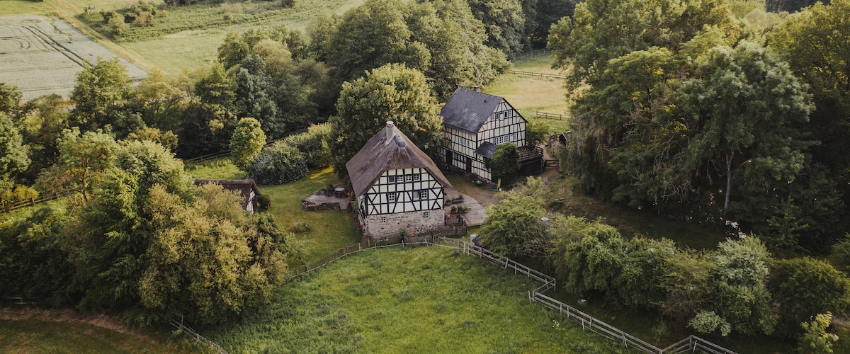 An aerial view reveals a historic farmhouse surrounded by lush green landscapes and trees. The building features traditional timber framing, set amidst vibrant vegetation and open fields, illustrating the natural beauty of the countryside.