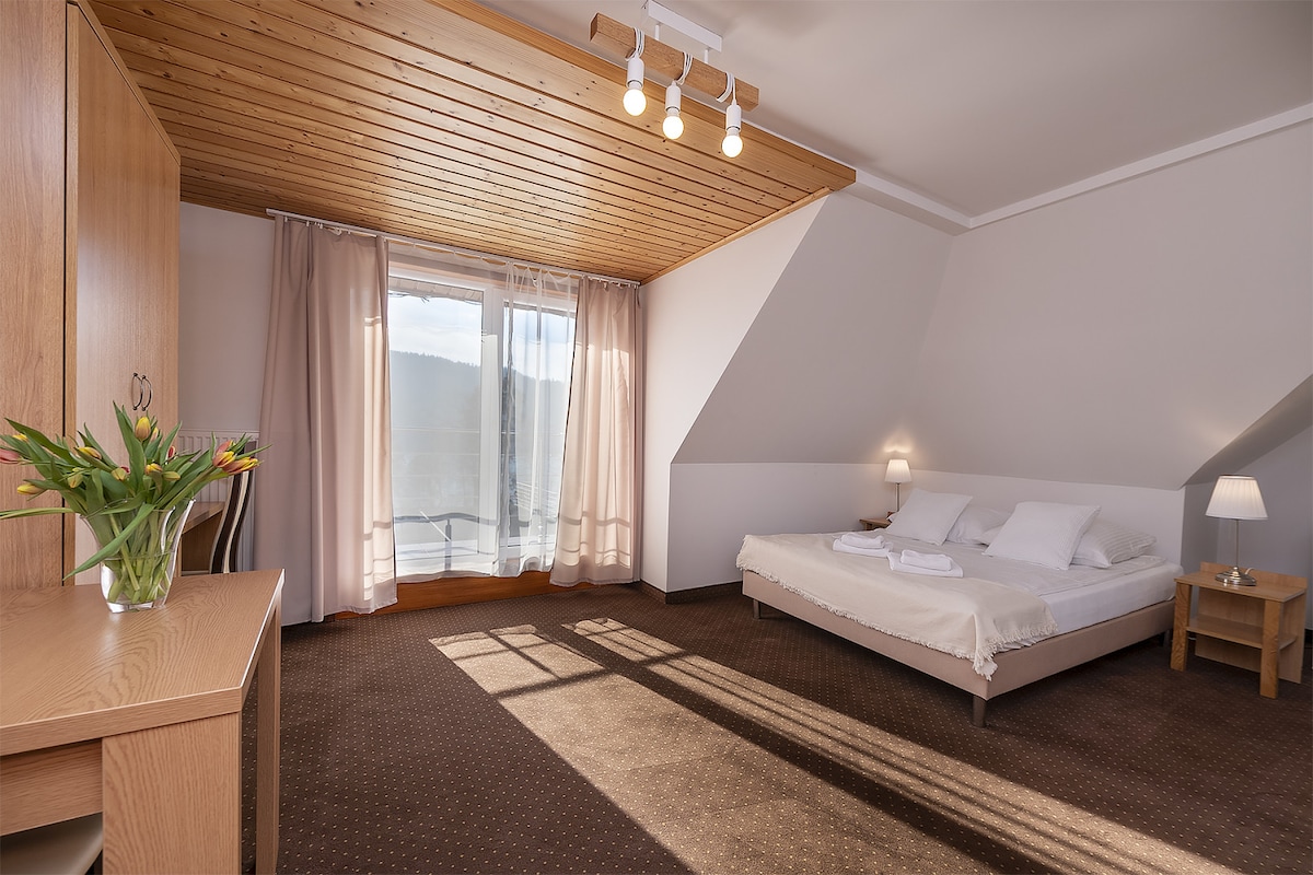 A spacious guest room features a light wood ceiling and a large window, allowing natural light to fill the space. The room includes a bed with crisp white linens, a simple bedside table, and a modern desk with a vase of fresh flowers.