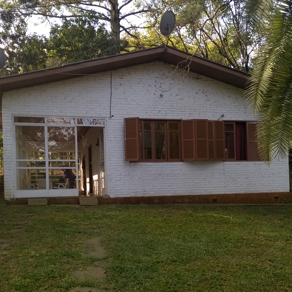 The exterior of a white, single-story house is visible, featuring large glass doors and wooden shutters. A well-maintained lawn surrounds the house, with trees providing shade. The simple, rustic design blends with the natural environment, showcasing a peaceful outdoor setting.