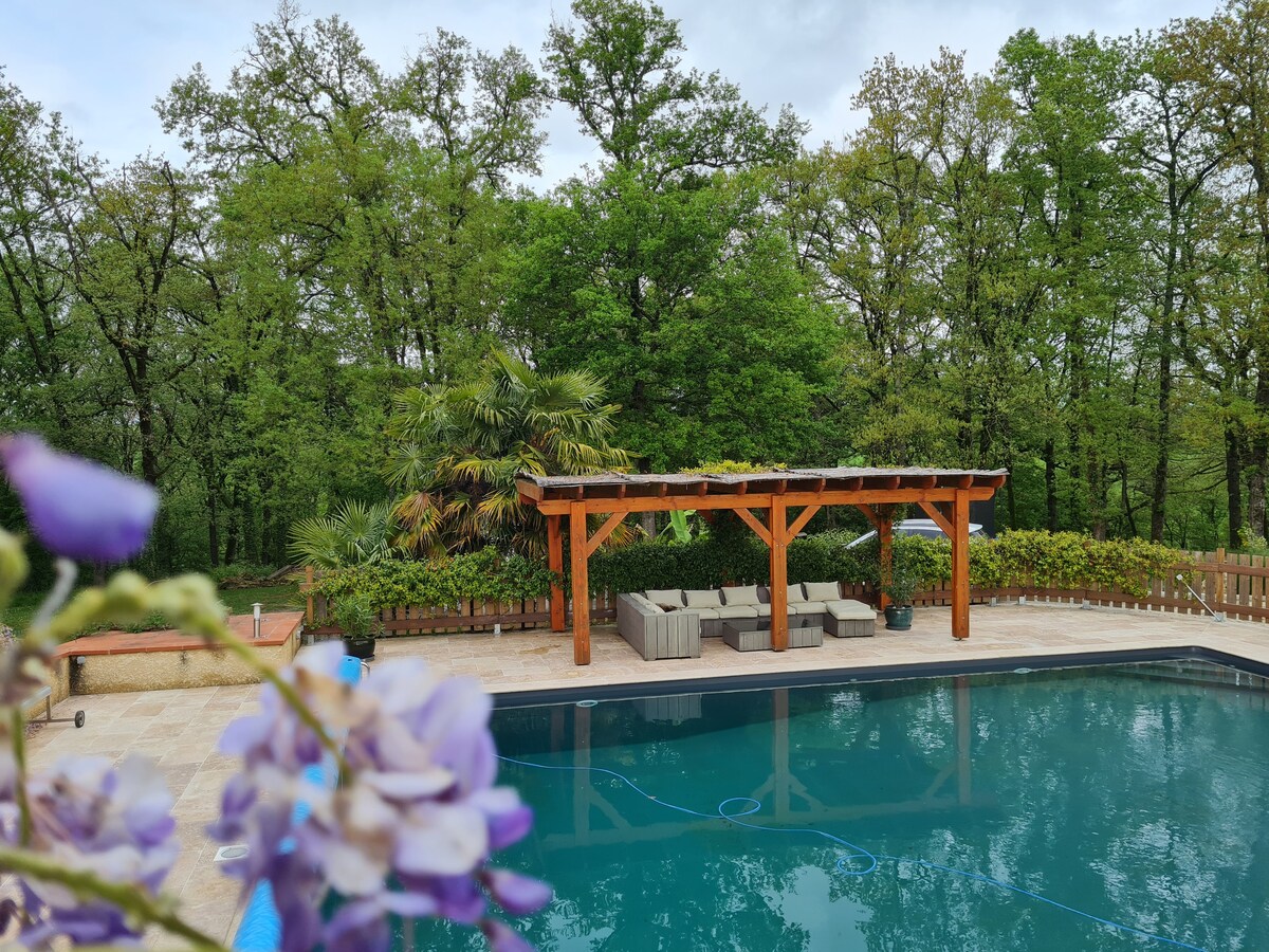The outdoor area features a refreshing pool surrounded by greenery, with a wooden pergola providing shaded seating. Lush trees and plants create a natural backdrop, enhancing the peaceful atmosphere of the setting.