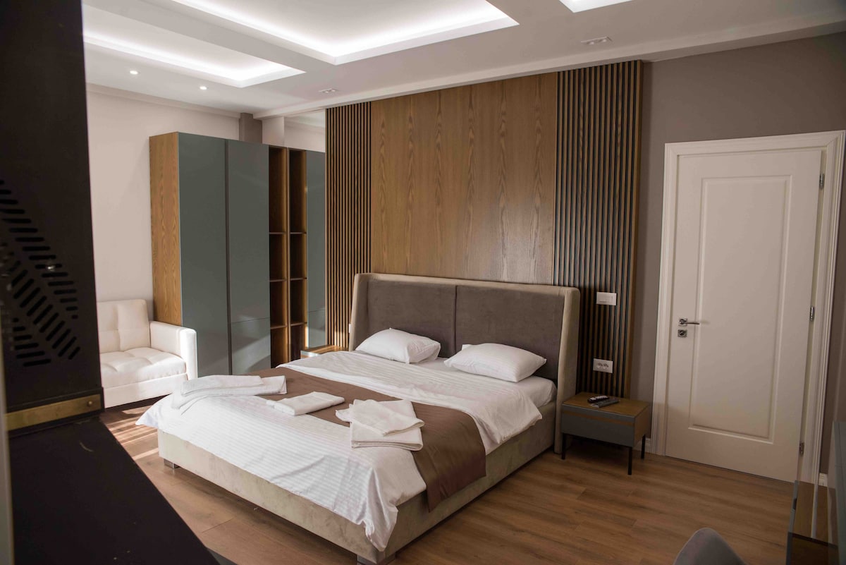 The bedroom features a spacious bed with neutral linens, complemented by a modern wooden accent wall. A bedside table and a comfortable seating area are visible. Natural light enters from overhead lighting, enhancing the calming atmosphere of the room.
