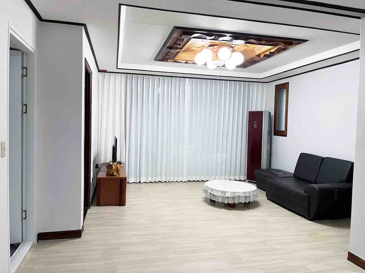 A spacious living area is presented with a comfortable black sofa and a circular coffee table. Light-colored flooring complements the white walls adorned with sheer curtains. A ceiling feature with multiple light fixtures adds to the room's ambiance, while a small entertainment unit is visible.