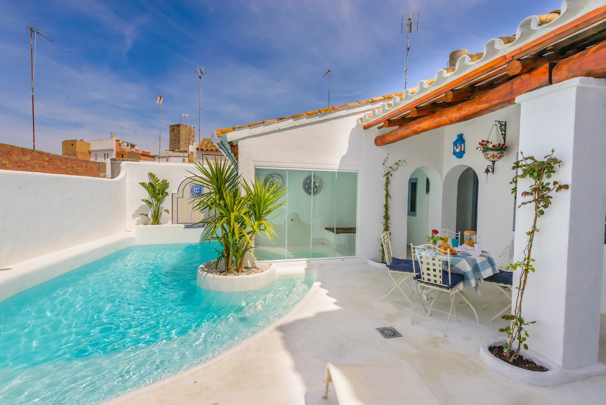 A tranquil area is highlighted with a curved swimming pool surrounded by white walls and a sunlit terrace. The outdoor space features a bistro-style table set for dining, along with decorative palms and a few seating options. Clear glass doors lead into the indoor area.