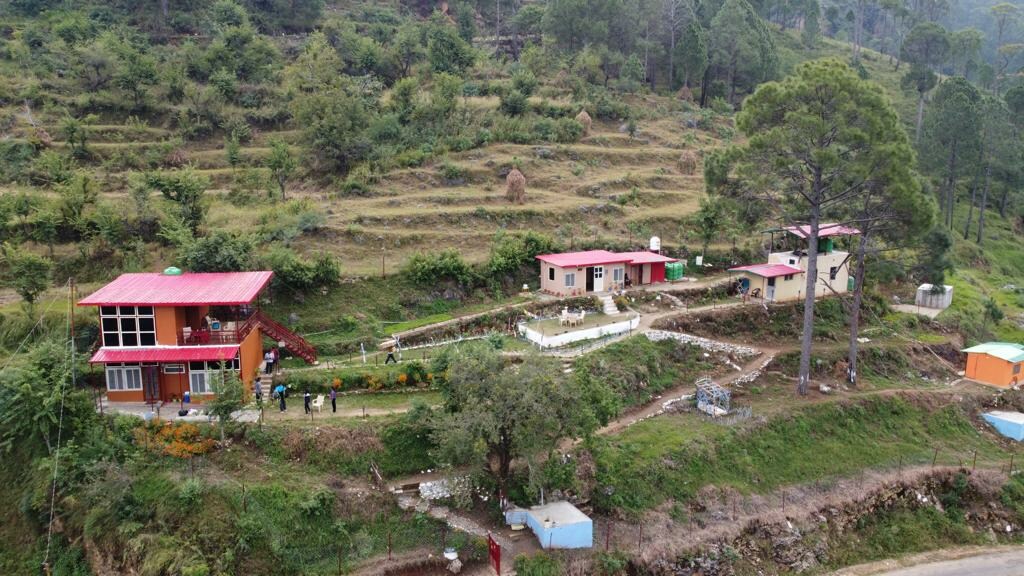 A serene landscape featuring multiple homestay buildings surrounded by lush greenery and trees. The vibrant red roofs contrast with the natural environment, while winding pathways connect the structures. The area is marked by cultivated terraces reflecting the local topography.