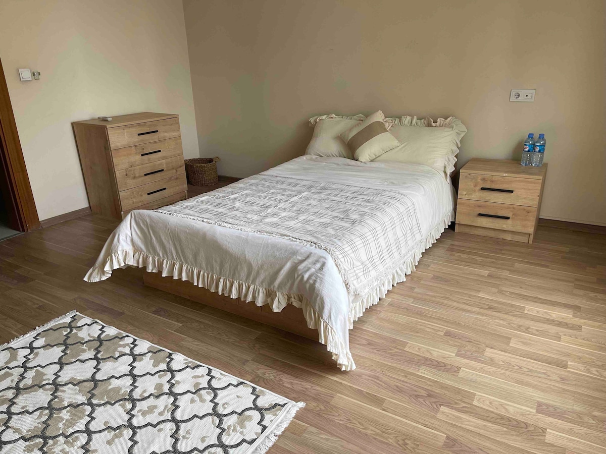 A comfortable sleeping area features a spacious bed with a light-colored blanket and decorative pillows. Two wooden nightstands are positioned on either side, and a small woven basket adds a touch of organization. A patterned rug is placed beneath the bed on the hardwood floor.