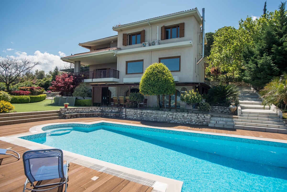 A two-story villa is set among lush greenery, featuring large windows that offer views of the surrounding landscape. A spacious swimming pool with inviting blue water is accompanied by wooden decking and chairs, creating a relaxing outdoor space.