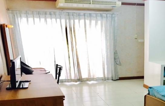 A bright room features large windows covered with sheer curtains, allowing ample natural light. A wooden desk and a television are positioned nearby, while a spacious bed is visible in the corner. Light-colored tiles enhance the open feel of the space.