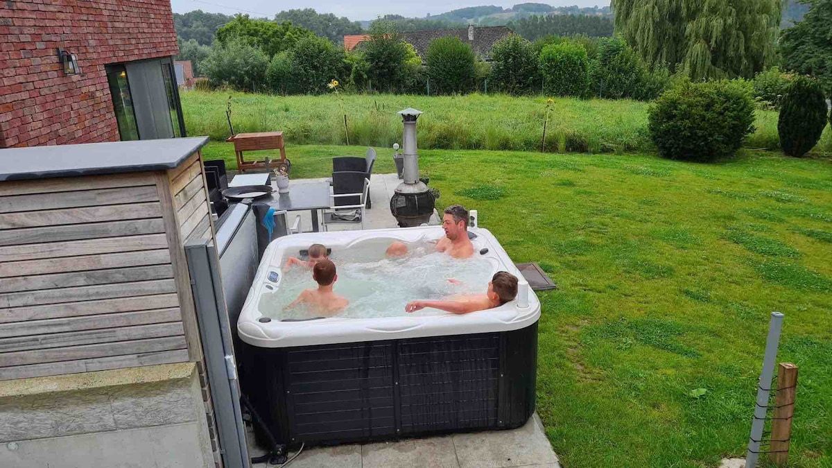 A hot tub is situated in a grassy area, surrounded by a patio. Several individuals are seen enjoying the warm water. In the background, a scenic view of rolling hills and greenery can be observed, creating a peaceful outdoor environment.