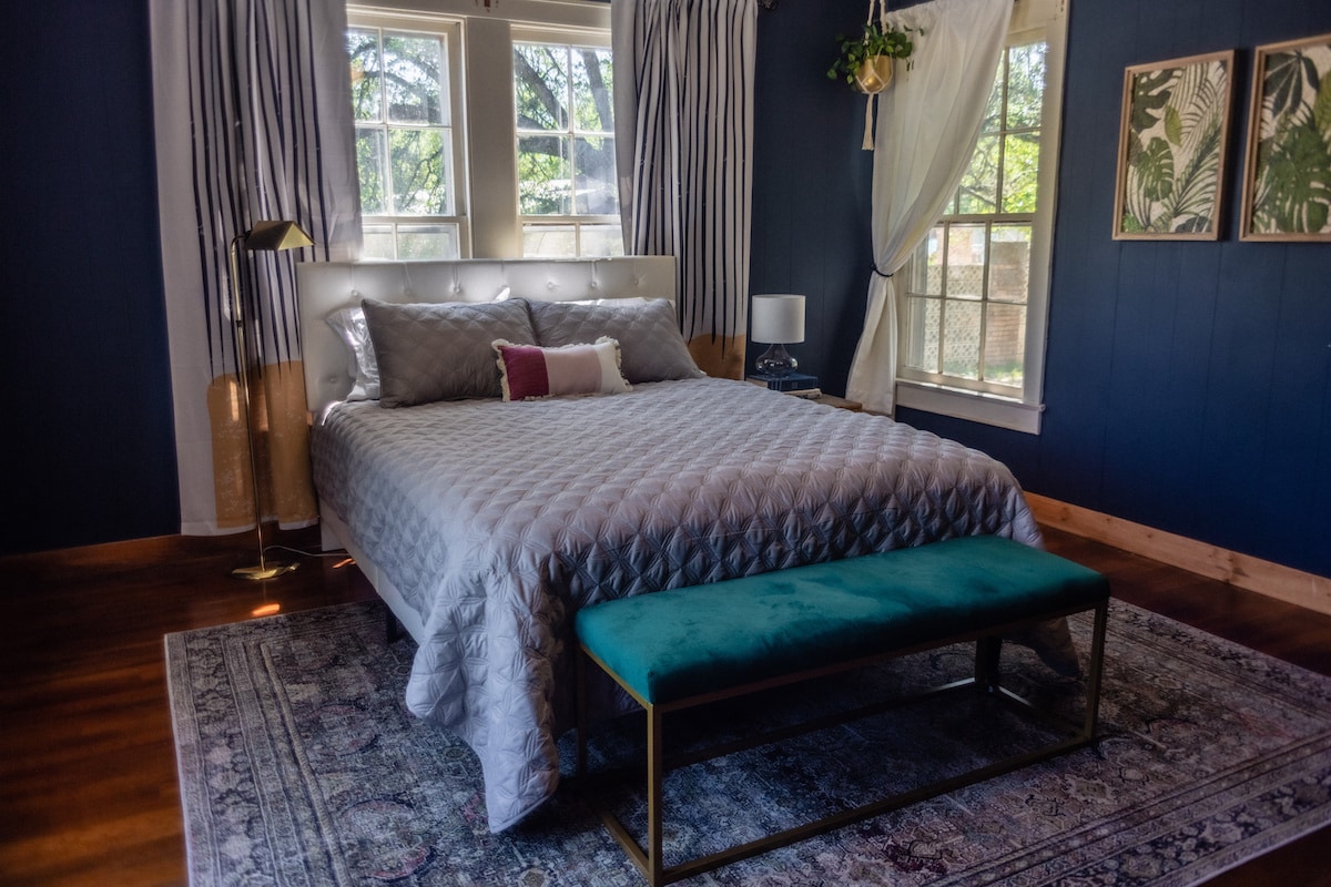 A spacious bedroom features a queen bed with a quilted coverlet, complemented by a decorative throw pillow. A velvet bench sits at the foot of the bed. Natural light streams through large windows adorned with patterned drapes, casting a warm glow on the hardwood floor.