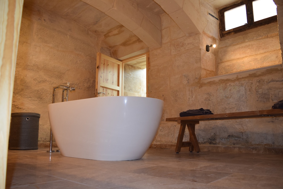 A spacious bathroom features a freestanding white bathtub positioned on neutral-toned tiled flooring. Original stone walls create a rustic ambiance, while a wooden bench adds warmth. Soft natural light enters through a small window, enhancing the tranquil environment of the luxury space.