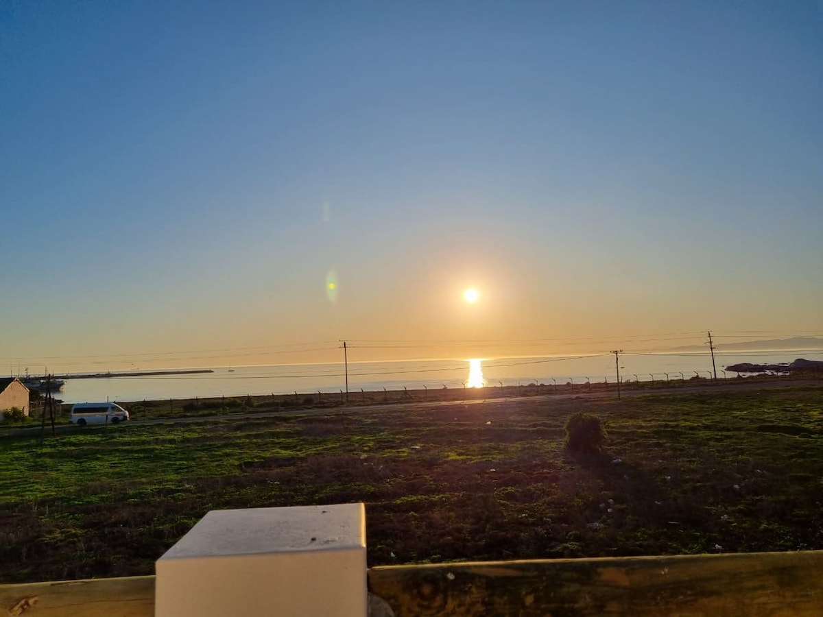 A serene view of a sunset over the calm waters, where the sun's reflection glistens on the surface. The horizon is clear, and gentle hills can be seen in the distance. An open area is visible in the foreground, enhancing the sense of tranquility.