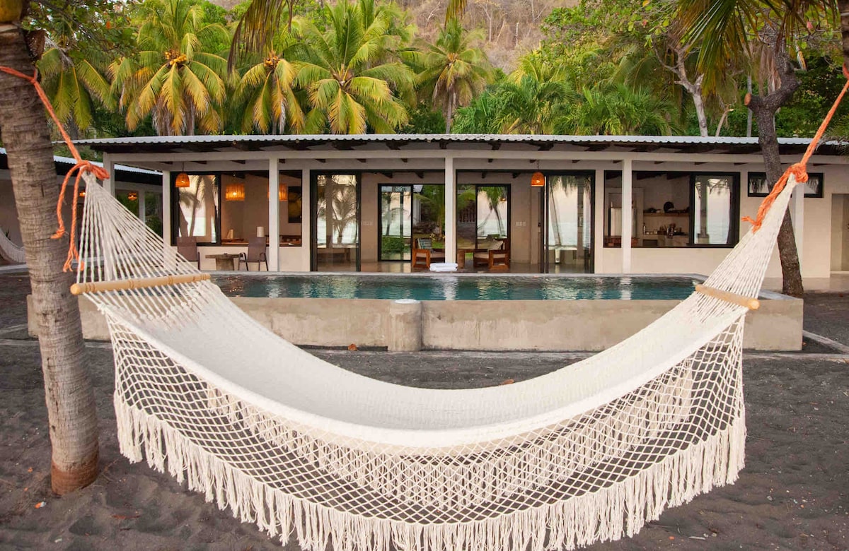 A spacious hammock is suspended between two palm trees, offering a relaxing spot near the pool area. Behind, a modern building features large glass doors that invite natural light. Lush palm trees frame the background, enhancing the serene outdoor setting.