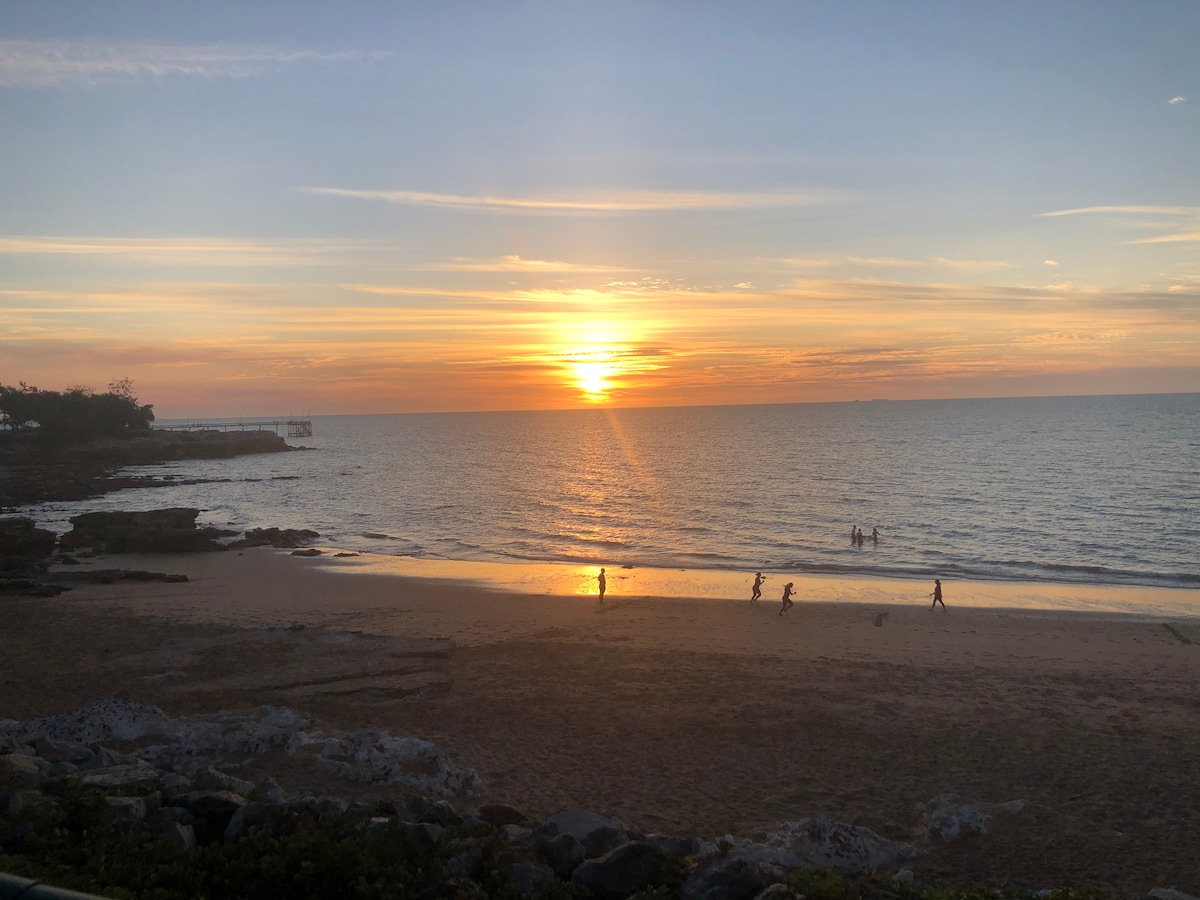 The image captures a serene beach scene at sunset, with soft waves lapping against the shore. Silhouettes of individuals can be seen walking along the water’s edge, while the golden sun dips below the horizon, casting warm hues over the landscape.