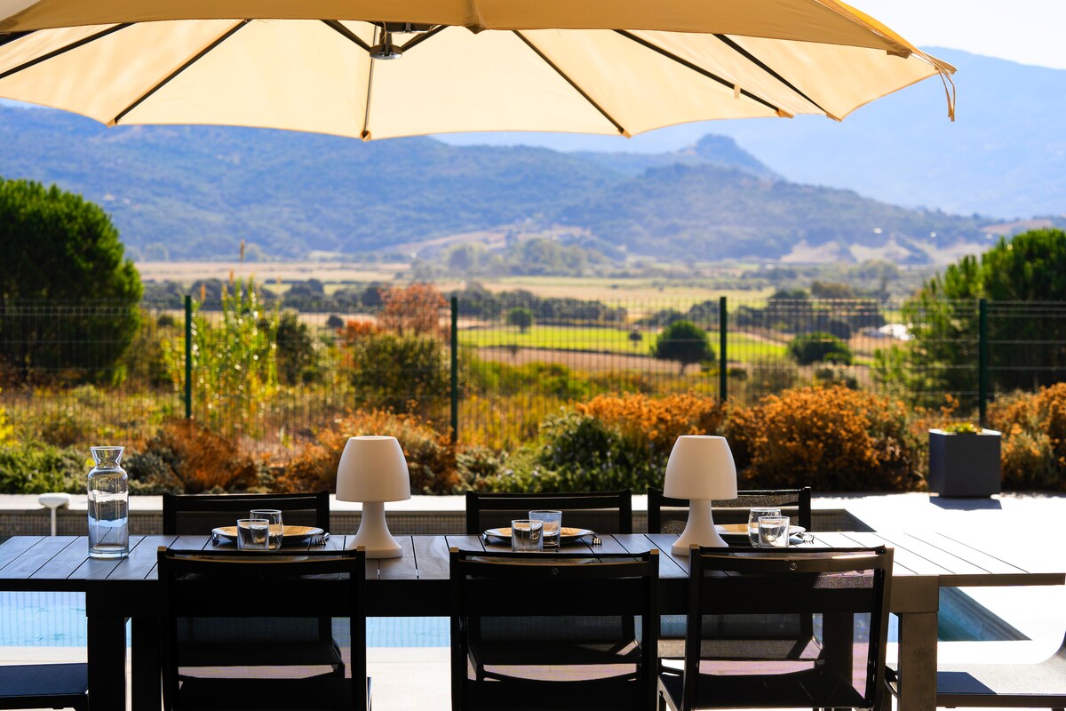 An outdoor dining table is set for a meal, featuring four chairs. A large umbrella provides shade, while a scenic view of rolling hills and greenery is visible in the background. The space offers a relaxing atmosphere for enjoying meals al fresco.