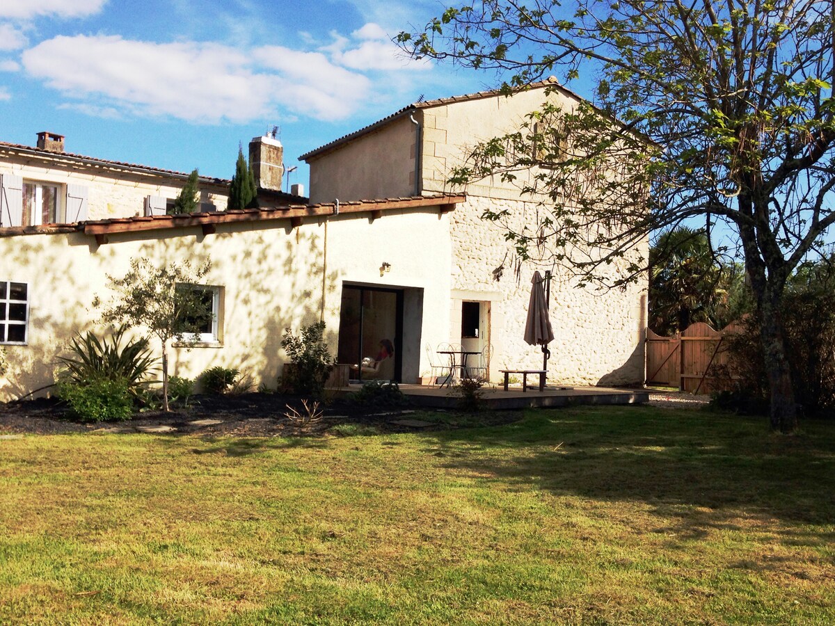 A charming stone house is depicted, featuring a grassy yard and a patio area. A large umbrella provides shade for a small table and chair set. Surrounding greenery includes a small tree and shrubs, with a bright sky above creating a serene outdoor environment.