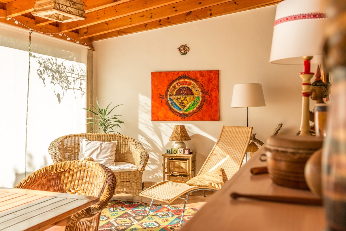 A sunlit relaxation area features a woven chair alongside a reclining lounge chair. A colorful rug is visible on the floor, while an artwork depicting a landscape is mounted on the wall. Greenery can be seen in the background, enhancing the peaceful ambiance.