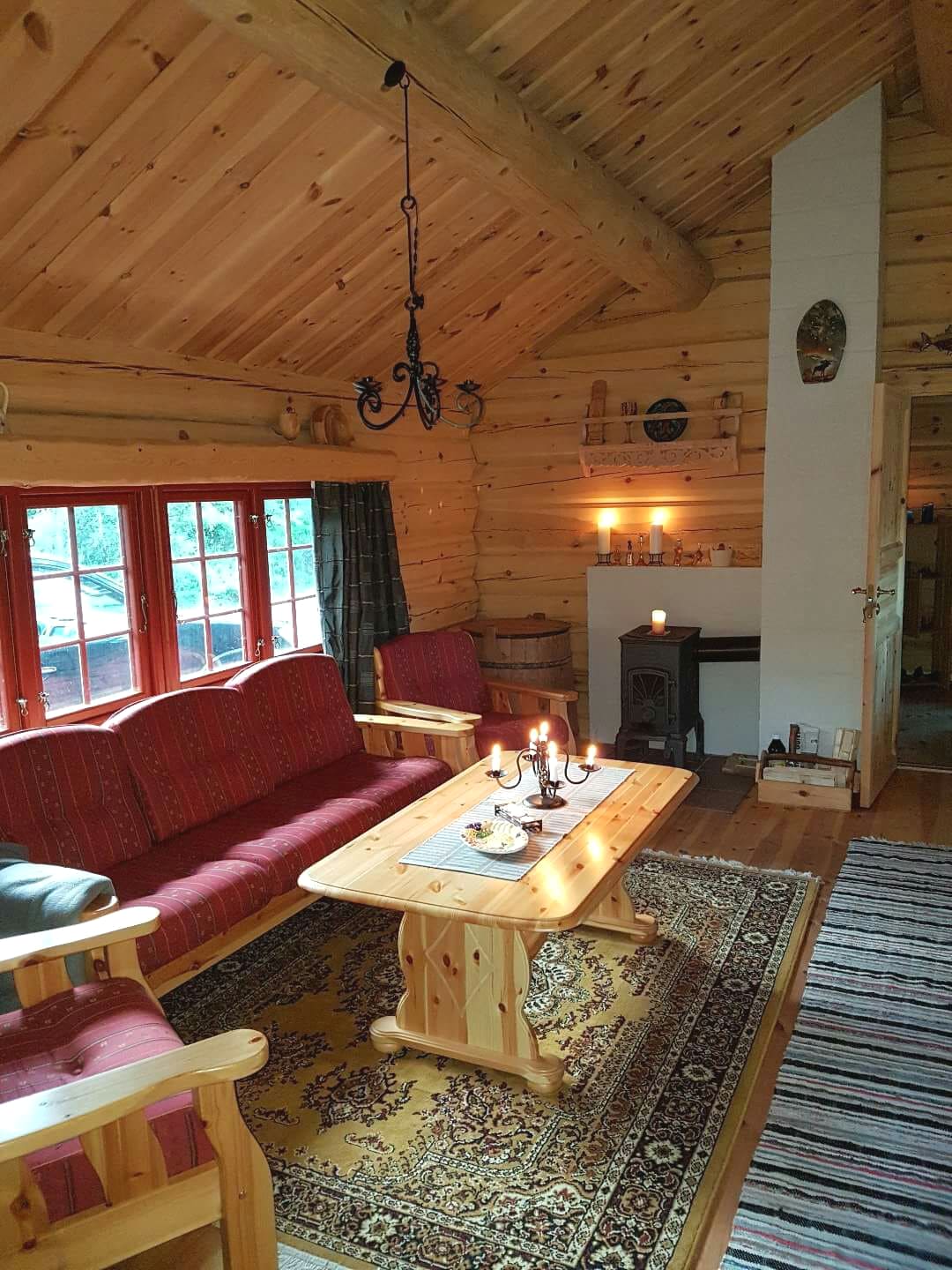 A cozy living area features wooden walls and ceiling, creating a warm atmosphere. Comfortable seating is arranged around a wooden table, with a decorative rug beneath. A wood-burning stove stands beside a lit candle, providing soft light. Large windows invite natural light while offering views of the surrounding woods.