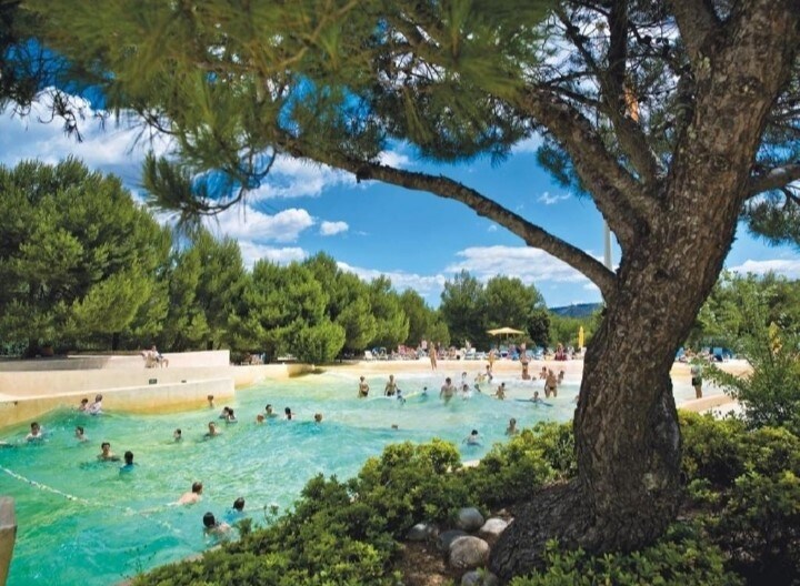 A pool area is surrounded by lush greenery and tall trees, providing shade for guests enjoying the water. The turquoise water reflects the blue sky, while several people are seen swimming and relaxing in the inviting space.