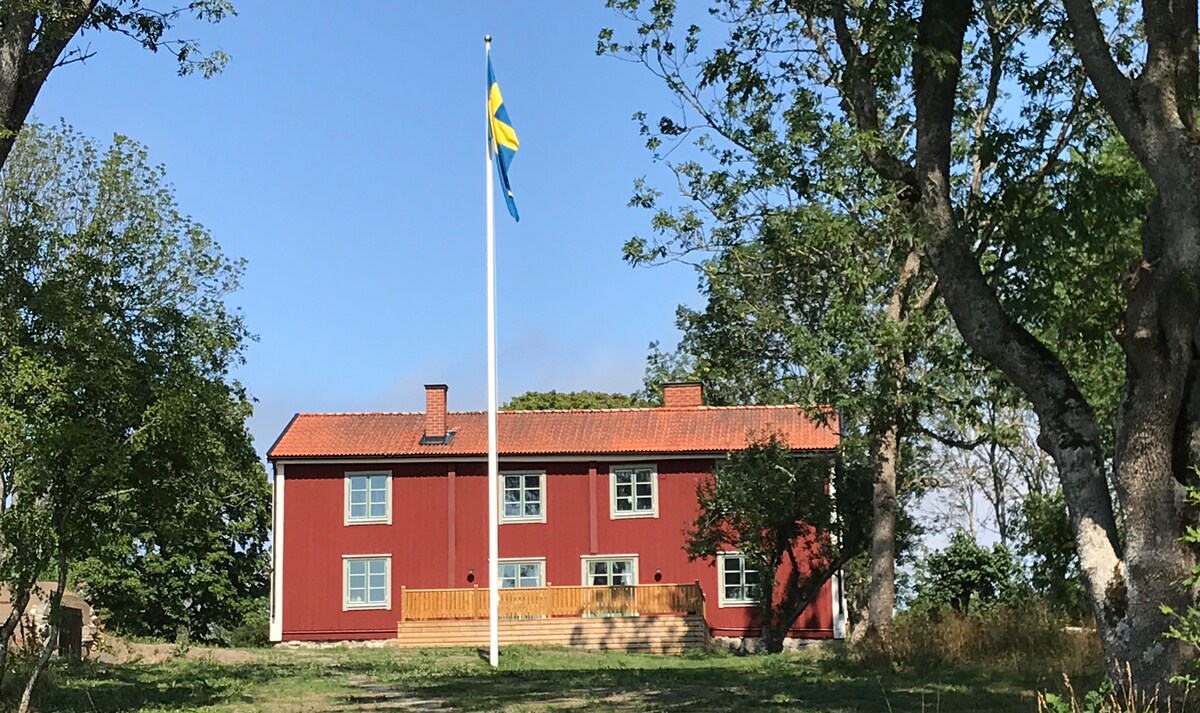 A charming red house is positioned on a hill, surrounded by trees. The building features a welcoming porch with steps leading up, and a flagpole displaying the Swedish flag stands prominently in front.