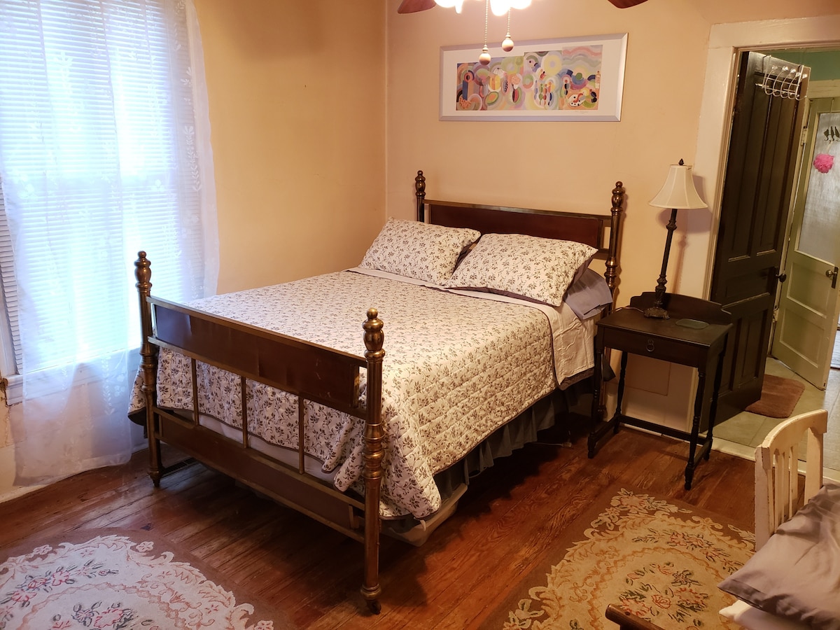A spacious bedroom features a vintage metal bed, dressed with a floral patterned bedspread. A bedside table stands next to the bed, complemented by a lamp. Light filters through a sheer-curtained window, and a charming area rug adorns the hardwood floor.
