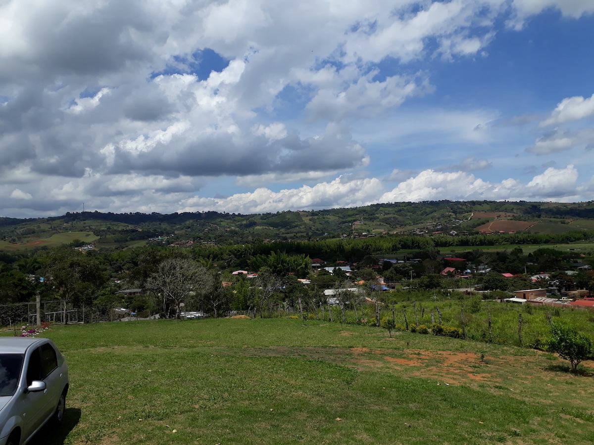 A scenic view of the surrounding landscape is captured, showcasing rolling hills, patches of greenery, and a distant village. The sky is partly cloudy, enhancing the natural beauty. A well-maintained grassy area is visible in the foreground, accompanied by a parked car.