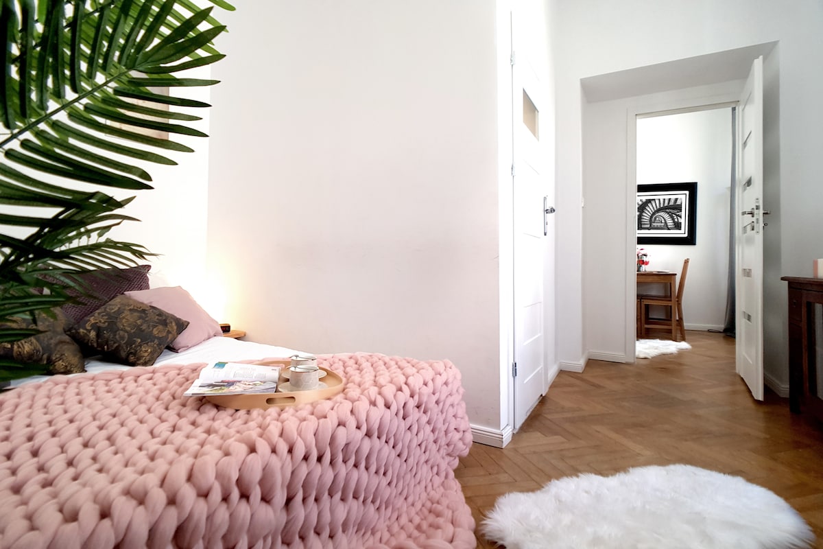 A bright and inviting sleeping area features a soft, textured pink blanket draped over a spacious bed. A vibrant plant adds a touch of greenery, while a doorway leads to a dining area in the background, visible through an open white door.
