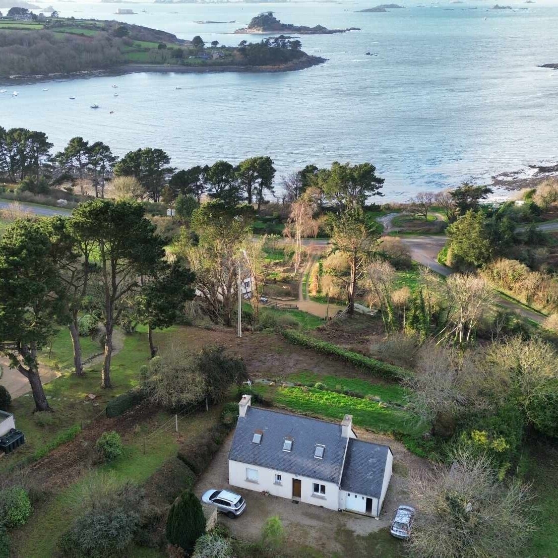 An aerial view showcases a charming house surrounded by lush greenery, leading to a serene coastline. The distant view reveals calm waters dotted with boats, while the garden and outdoor space are clearly defined, offering a peaceful retreat.