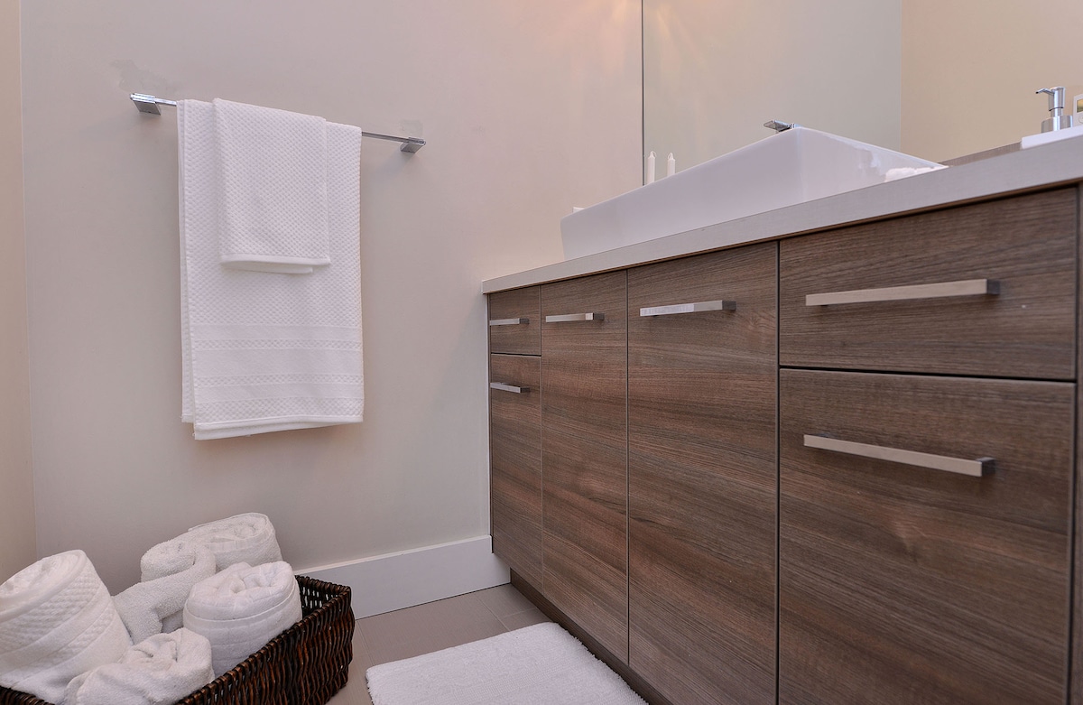 A modern bathroom features a sleek vanity with wooden cabinetry and a minimalist sink. Fresh white towels are neatly hung and stacked, offering convenience. A soft bath mat is positioned on the floor, enhancing comfort in the space.