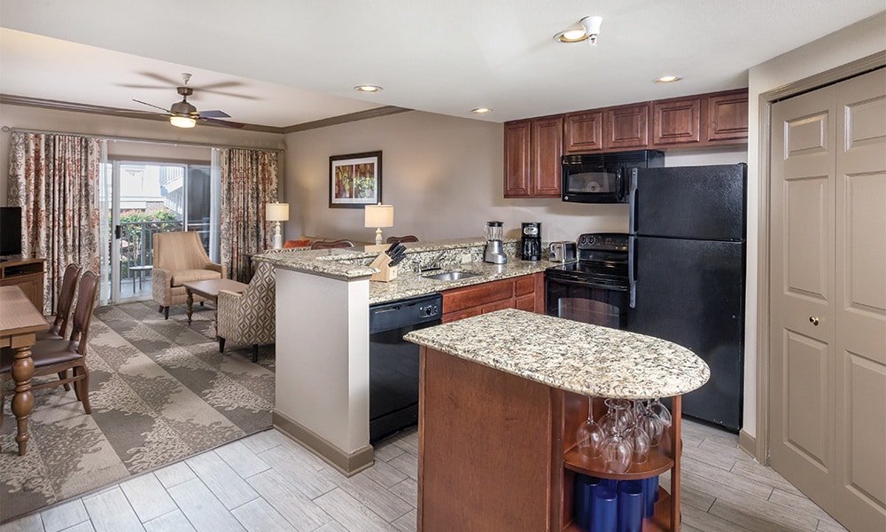 A modern kitchen with granite countertops is showcased alongside dark wood cabinetry and stainless steel appliances. The space includes a dining area with wooden chairs and a small table. Natural light enters through a sliding glass door, which leads to a patio area.