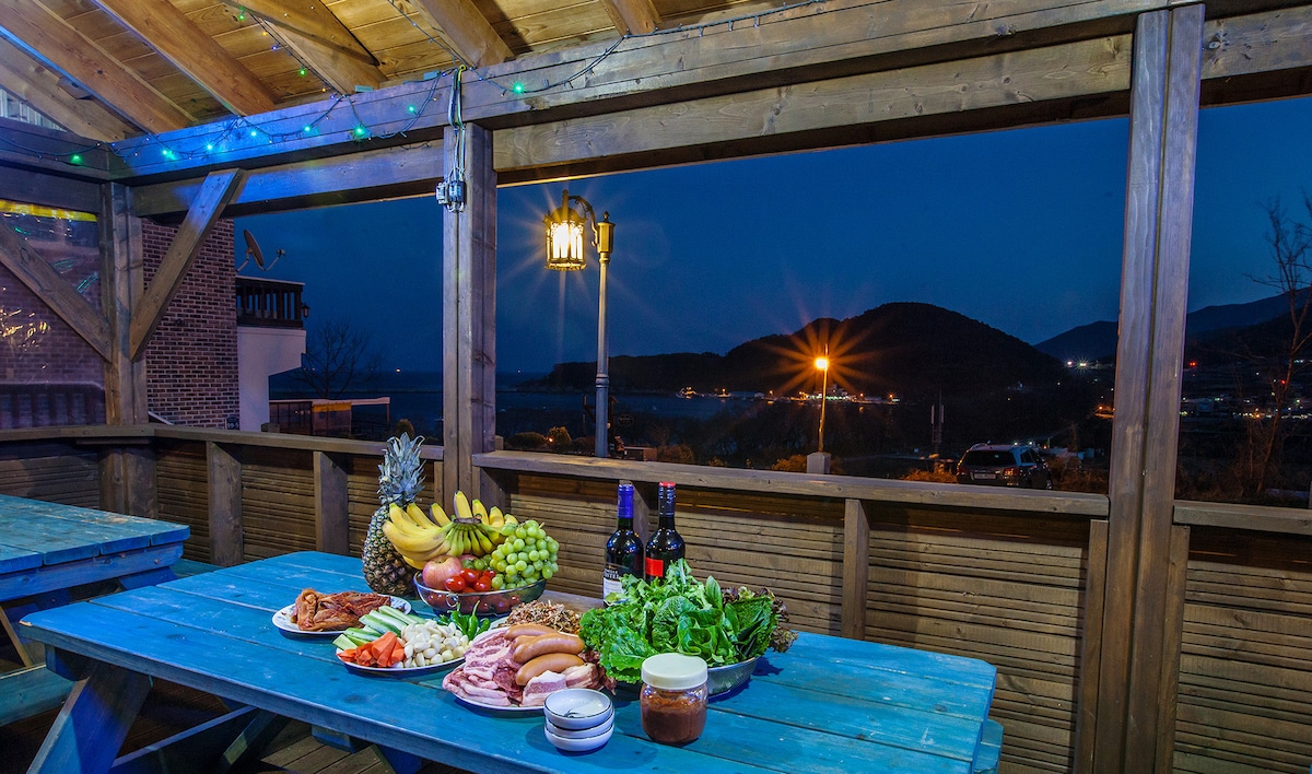 An outdoor dining area is illuminated by soft lighting, featuring a large blue table set with various foods including fruits, meats, and vegetables. A scenic view of the surrounding hills and a bay is visible in the background, enhancing the inviting atmosphere.