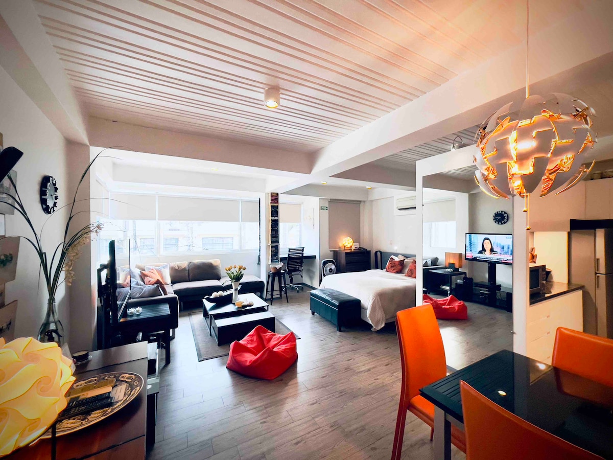The spacious studio apartment features a layout that includes a large bed, a sofa area, and a dining table. Natural light fills the space through large windows, while colorful bean bags add a casual touch. A sleek pendant light hangs above the dining area.