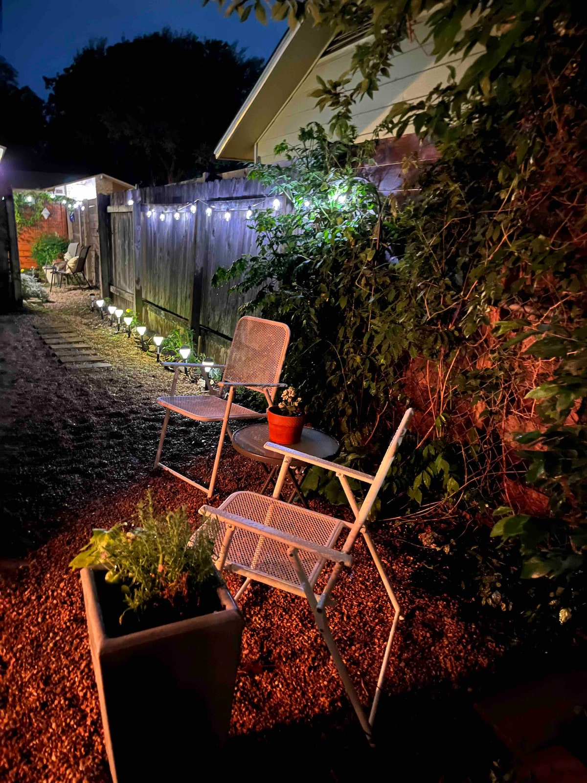 An outdoor sitting area is illuminated by soft string lights, featuring two metal chairs beside a potted plant. A gravel path leads through a lush garden, accented by green foliage and ambient lighting in the evening setting.