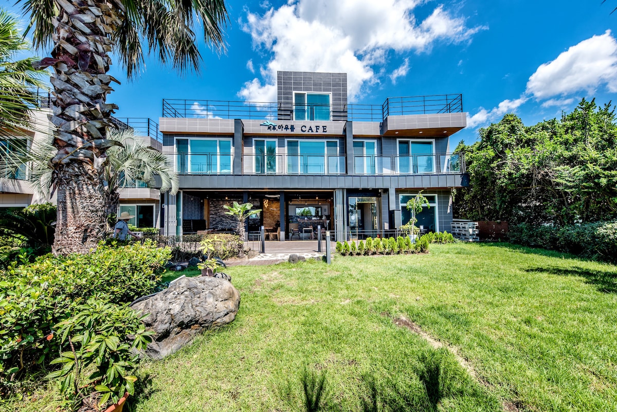 A spacious two-story building features large glass windows and a modern exterior, surrounded by well-maintained green lawns and palm trees. A patio area with outdoor seating is visible, creating a serene environment. The cafe's name is displayed prominently above the entrance.