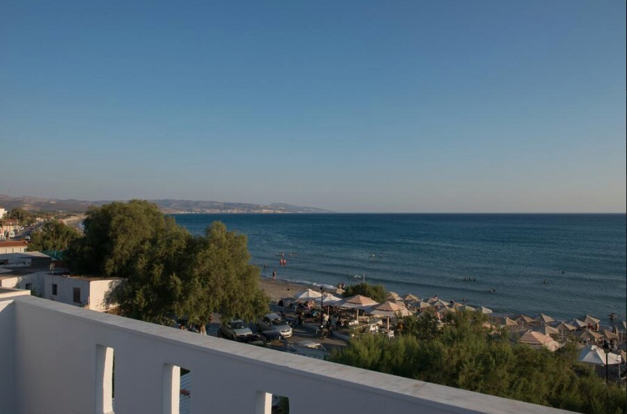 A scenic view from the balcony showcases the sandy beach and calm blue waters of the sea. Beach umbrellas and sun loungers are visible along the shoreline, while distant hills are framed against the horizon. The clear sky complements the tranquil atmosphere.