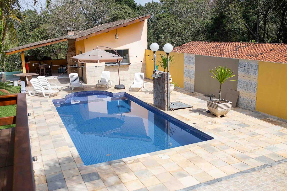 A modern outdoor area features a rectangular swimming pool surrounded by a stone patio. Multiple lounge chairs are positioned nearby, with a large umbrella providing shade. A stone wall adds texture, and a small plant sits in a decorative pot, enhancing the natural setting.