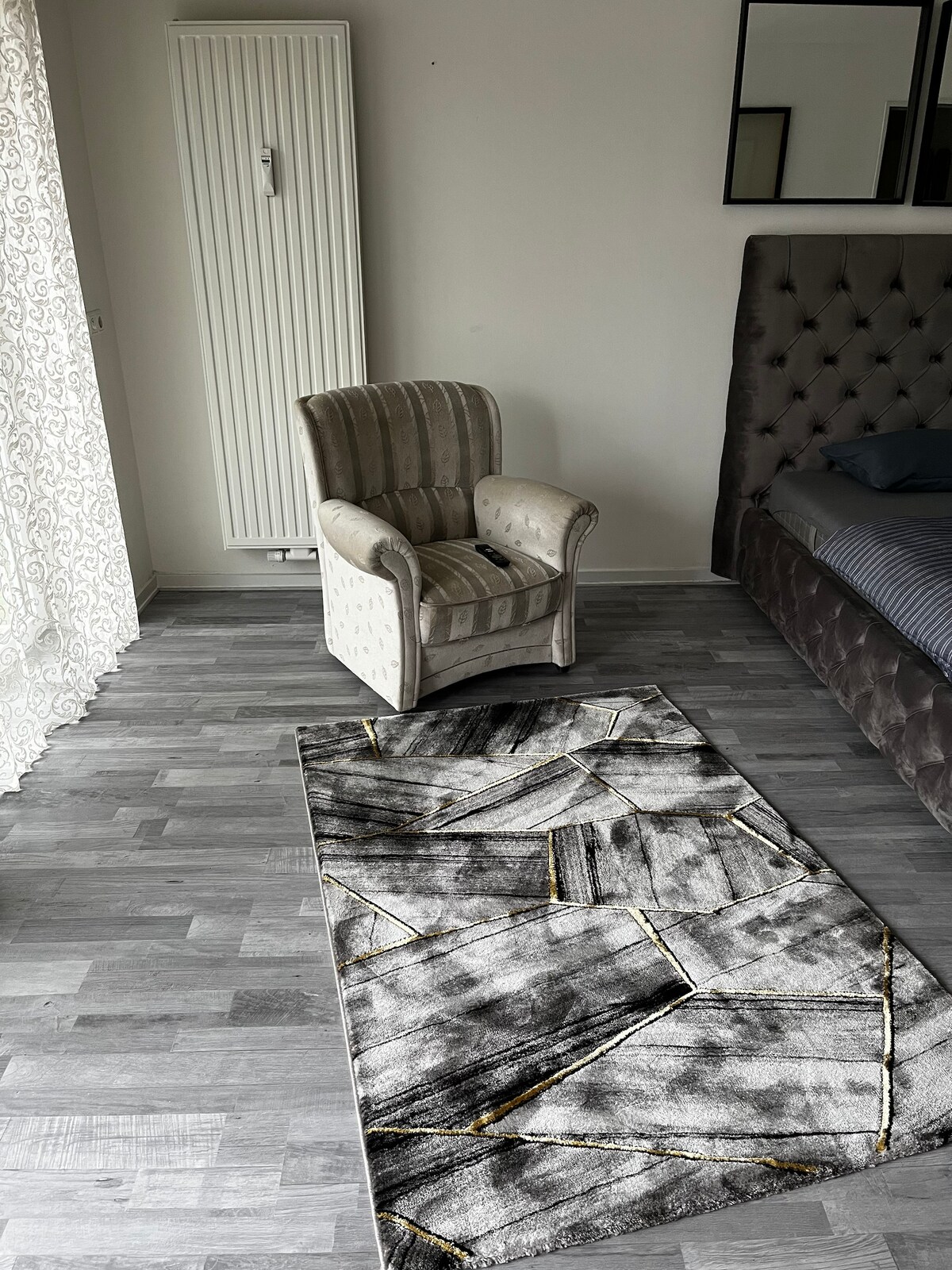 A cozy corner of the room features a plush armchair with a vertical striped pattern. The floor is adorned with a geometric area rug in monochrome tones, accented by gold lines. Natural light filters through sheer curtains, highlighting the room's subtle decor.
