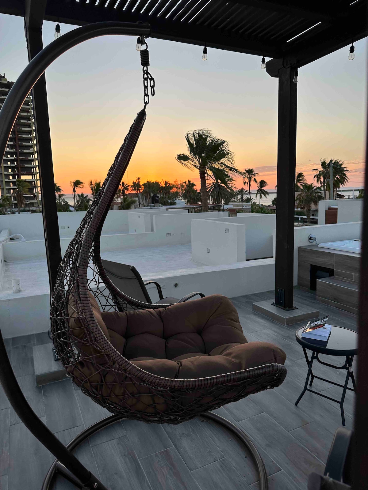 A hanging wicker chair is situated on a private terrace, facing a sunset with silhouettes of palm trees and buildings in the background. A small table beside the chair holds a cup, enhancing the relaxed environment. Soft lighting adds to the tranquil setting.